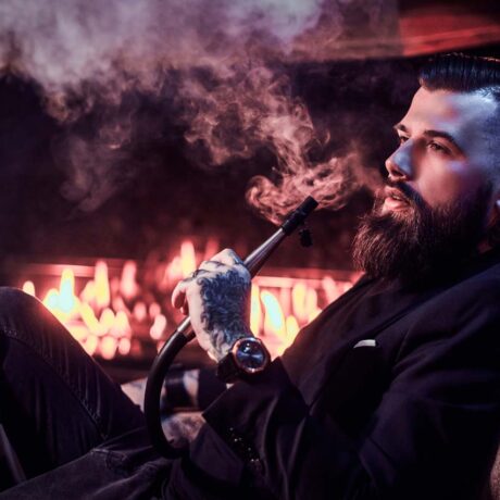 Portrait of tattooed bearded man which is smoking hookah, making nice vapour while chilling on the armchair near fire place.